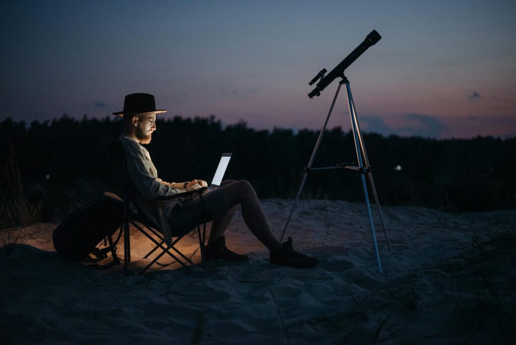 Remote Work Freedom: Purpose-Driven Life Beyond the 9-to-5 Man working remotely on a laptop in nature at dusk with a telescope beside him, symbolizing purpose-driven freedom from the 9-to-5.
