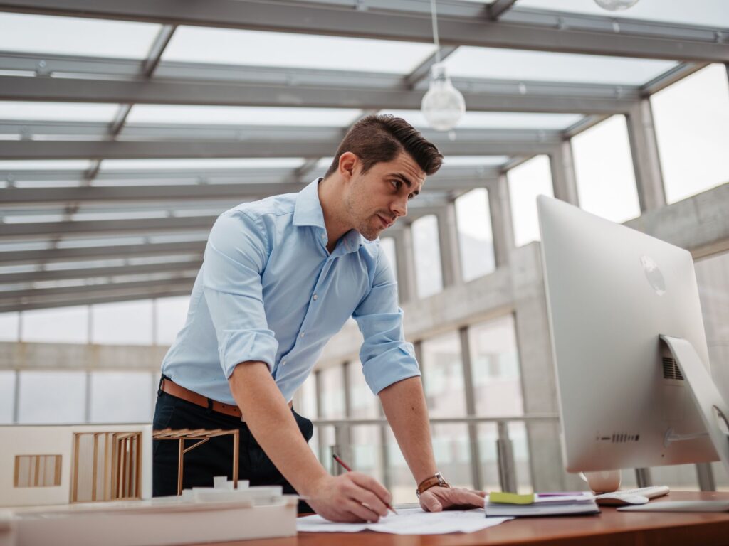 confident-early-career-engineer Confident young professional standing at a desk, focusing on work in a modern office with large windows and a computer monitor.